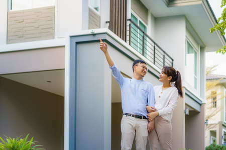 Portrait of Asian couple walking hugging and pointing together looking happy in front of their new house to start new life. Family, age, home, real estate and people concept.の写真素材
