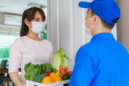 Asian woman costumer wearing face mask and glove receive groceries box of food, fruit, vegetable and drink from delivery man   in front of the house during time of home isolation.の写真素材