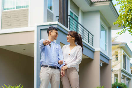 Portrait of Asian young couple standing and hugging together and holding house key looking happy in front of their new house to start new life. Family, age, home, real estate and people concept.の写真素材