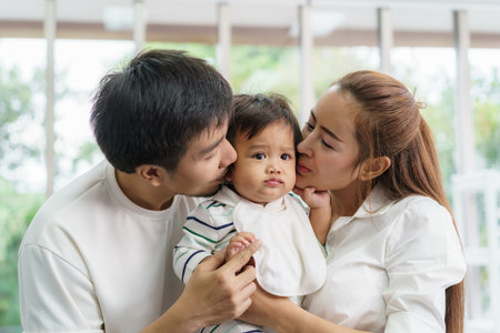 Asian family happy kiss little baby boy in living room at home, young parent and children enjoy love hug standing together.の写真素材