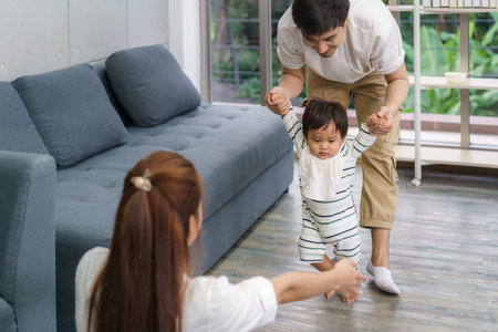 Asian son baby taking first steps walk forward to his mother. Happy little baby learning to walk with father help and teaching how to walk gentlyat homeの写真素材