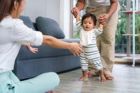 Asian son baby taking first steps walk forward to his mother. Happy little baby learning to walk with father help and teaching how to walk gentlyat homeの写真素材