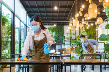 Portrait attractive Asian restaurant employee wear face mask and face shield cleaning table and floor with alcohol and wet wipe before welcome customer.の写真素材