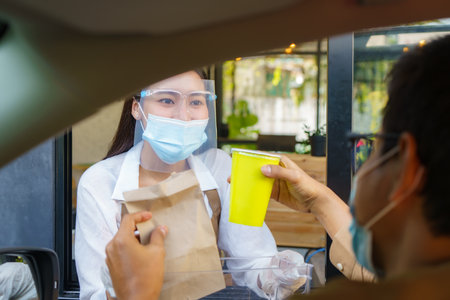Asian man in protective mask taking food bag and coffee with woman waitress wearing face mask and face shield at drive thru during coronavirus outbreak.の写真素材
