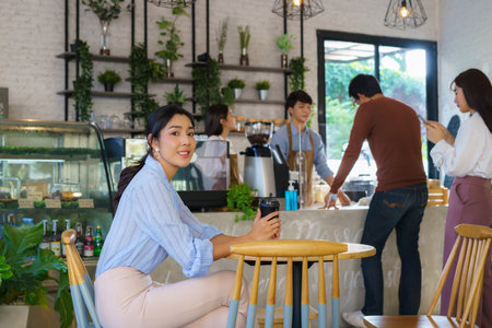Asian woman sitting in cafe and feeling happy while she drink coffee and another people queue order beverage with barista .の写真素材