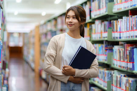 Portrait of Smart Asian woman university student reading book and looking at camera between bookshelves in campus library with copyspace.の写真素材