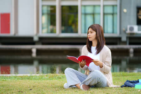 Portrait of Asian woman university student aitting on grass in campus looking happy and reading a book in park.の写真素材
