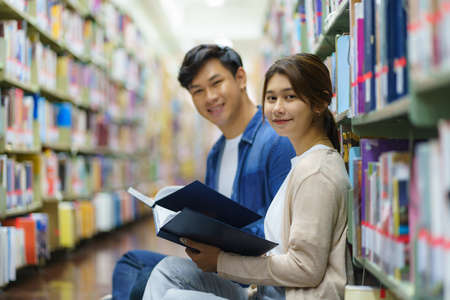Portrait of Smart Asian man and woman university student reading book together and. looking at camera between bookshelves in campus library with copyspace.の写真素材