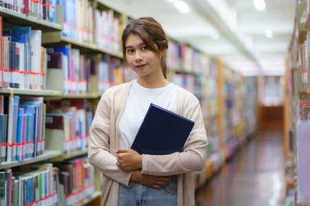 Portrait of Smart Asian woman university student reading book and looking at camera between bookshelves in campus library with copyspace.の写真素材