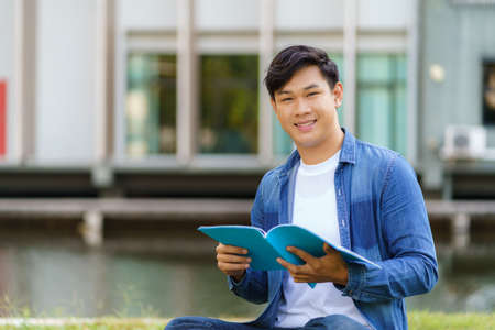 Portrait of Asian man university student aitting on grass in campus looking at camera and smile and reading a book in park.の写真素材