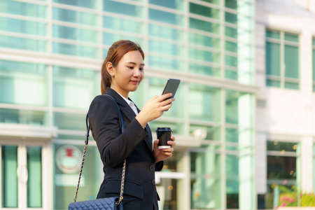 Asian executive working woman holding coffee cup and using a mobile phone in the street with office buildings in the background in Bangkok, Thailand.の写真素材