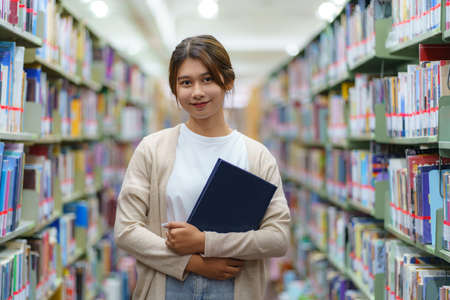 Portrait of Smart Asian woman university student reading book and looking at camera between bookshelves in campus library with copyspace.の写真素材