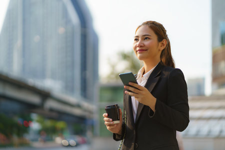 Asian executive working woman holding coffee cup and using a mobile phone in the street with office buildings in the background in Bangkok, Thailand.の写真素材