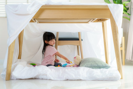 Asian cute little girl play cooking and feeding food to her doll while sitting in a blanket fort in living room at home for perfect hideout away from their other family members and for them to play imaginatively.の写真素材