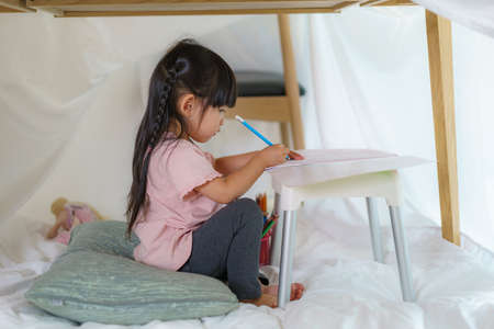 Asian cute little girl drawing in paper while lying in a blanket fort in living room at home for perfect hideout away from their other family members and for them to play imaginatively.の写真素材