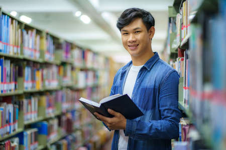 Portrait of Smart Asian man university student reading book and looking at camera between bookshelves in campus library with copyspace.の写真素材