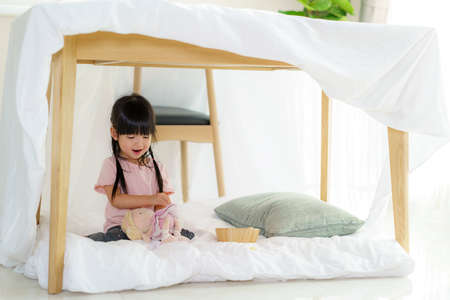Asian cute little girl play her doll while sitting in a blanket fort in living room at home for perfect hideout away from their other family members and for them to play imaginatively.の写真素材