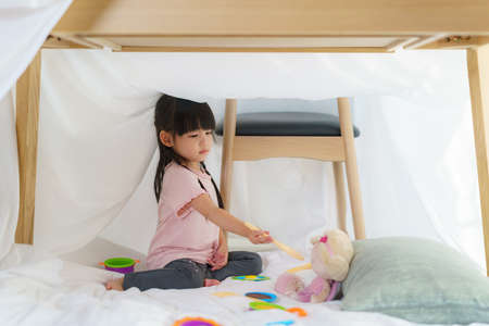 Asian cute little girl play cooking and feeding food to her doll while sitting in a blanket fort in living room at home for perfect hideout away from their other family members and for them to play imaginatively.の写真素材