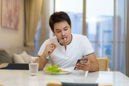 An Asian man is eating an American breakfast while sitting at chair to using phone, checking emails or news in the living room at home.の写真素材