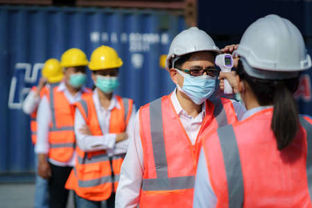 Asian woman foreman scanning fever temperature with digital thermometer to her staff worker wearing hygiene face mask protectの写真素材