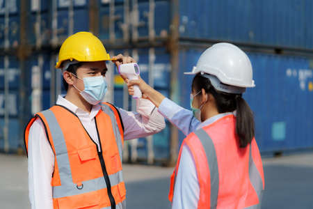 Asian woman foreman scanning fever temperature with digital thermometer to her staff worker wearing hygiene face mask protectの写真素材