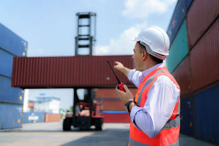 Caucasian foreman man is using walkie talkie to command his workers with crane lifting container in background in container depot terminal.の写真素材