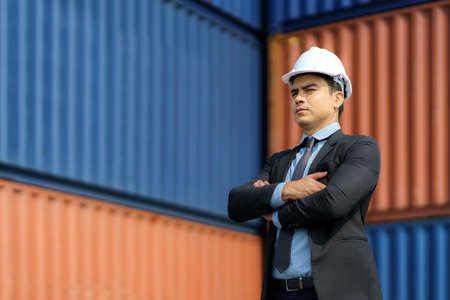 A Caucasian businessman is standing up and arm crossed to look at containers, planning a future, investing in a shipping business with containers in background.の写真素材