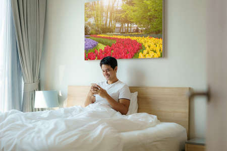 An Asian man sits in bed with a white blanket and checking his email on his cell phone in the morning at home before taking a shower.の写真素材