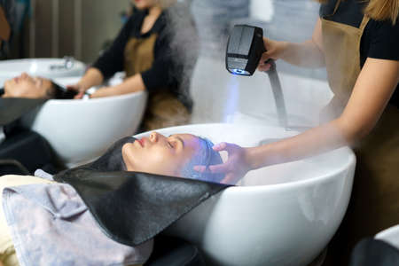 Hairdresser is using Nano Mist Hair Treatment Streamer Machine, Modern equipment for Young Happy Asian Beautiful Caucasian Woman Customer on Shampoo Chair In Hair Salon. Beauty Concept.の写真素材