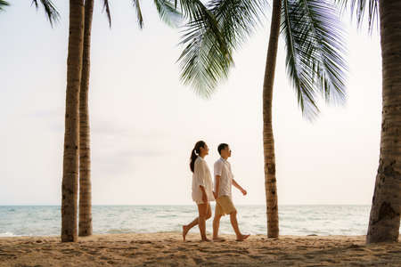 Asian couple is walking and holding hands on a beachfront beach sea with coconut trees while on vacation in the summer in Thailand.の写真素材