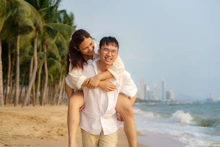 Asian man carry her girlfriend on his back and have fun on a beach with coconut palms while relaxing on a summer vacation.の写真素材