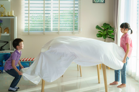 Asian siblings sister and brother stretching a white blanket over table furniture to make a fort in living room at home for perfect hideout away from their other family members and for them to play imaginatively.の写真素材
