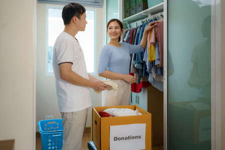 Asian couple are standing near closet of clothes in the dressing room and discussing clothing to donate for take to the donation box.の写真素材