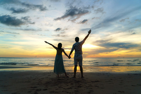 Romantic Asian couple standing and holding hand enjoying beautiful sunset on the beach during travel vacation holidays.の写真素材