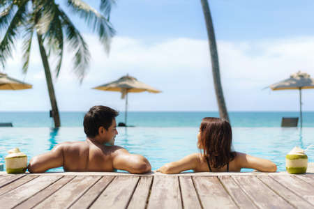Summer travel vacation concept, traveler asian couple  with coconut relax in luxury infinity pool hotel resort with sea beach and palm tree backgroundの写真素材