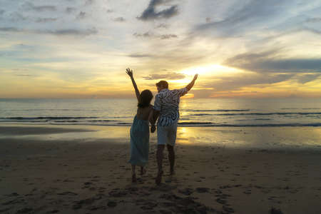Romantic Asian couple standing and holding hand enjoying beautiful sunset on the beach during travel vacation holidays.の写真素材
