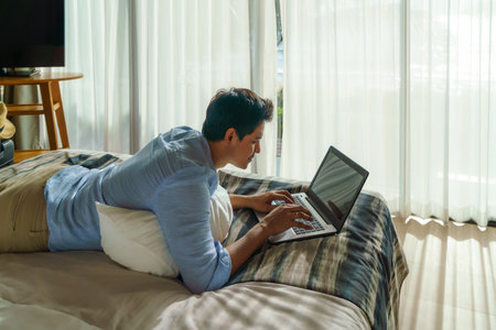 Young Asian businessman lying in bed and working with laptop in room at resort near sea during a summer vacation holiday travel.の写真素材