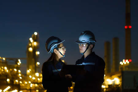 Asian engineers, man and woman are checking the maintenance of the oil refinery factory at night via...