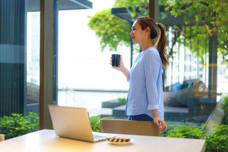 Asian woman drinking coffee in mug and smiling happily while taking a break from work at her...