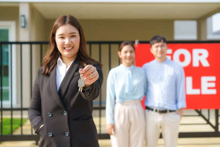 Asian real estate agent or realtor woman smiling and holding red file with showing house key with happy Asian couple hugging in front of thier house.の写真素材