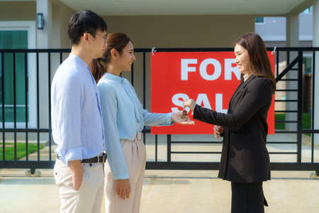 Asian happy smile young couple take keys new big house from real estate agent or realtor in front of their house after signing contract agreement, concept for real estateの写真素材