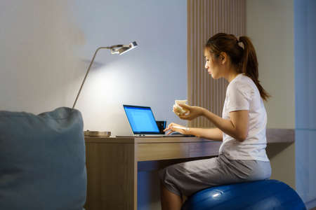 Asian woman relaxing on yoga ball and eating sandwich and chatting online with team of colleagues on laptop while work from home at home at night.の写真素材