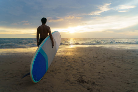 Asian man holding a sup board and paddle and walking on the beach. In the background, the ocean and the sunset. Back view. Summer surfing.の写真素材
