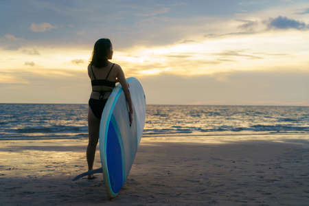 Asian woman holding a sup board and paddle and walking on the beach. In the background, the ocean and the sunset. Back view. Summer surfing.の写真素材