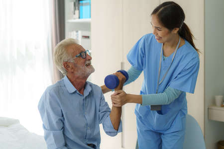 Senior man physiotherapy exercises with Asian woman caregiver or nurse physiotherapist in uniform helping aged male patient at home.の写真素材