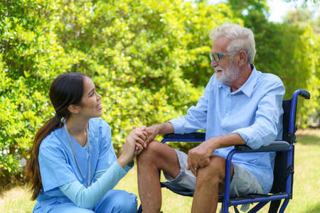 Asian nurse sitting on a hospital bed next to an older man helping hands care in garden at home....