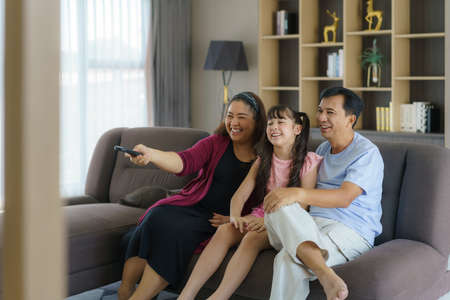 Happy Asian family with father, mother and daughter enjoy weekend activity spending more time at home and therefore probably more time in front of the televisionの写真素材