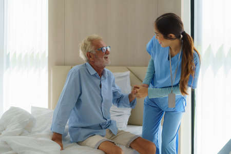 Asian nurse standing on a home bed next to an older man helping hands, care. Elderly patient care and health lifestyle, medical concept.の写真素材