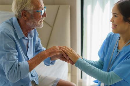 Asian nurse standing on a home bed next to an older man helping hands, care. Elderly patient care and health lifestyle, medical concept.の写真素材