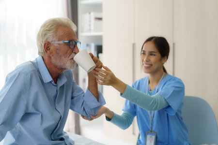 Asian elderly caregiver woman sitting on a hospital bed next to an older man helping drink a glass of water from the bedroom at home.の写真素材